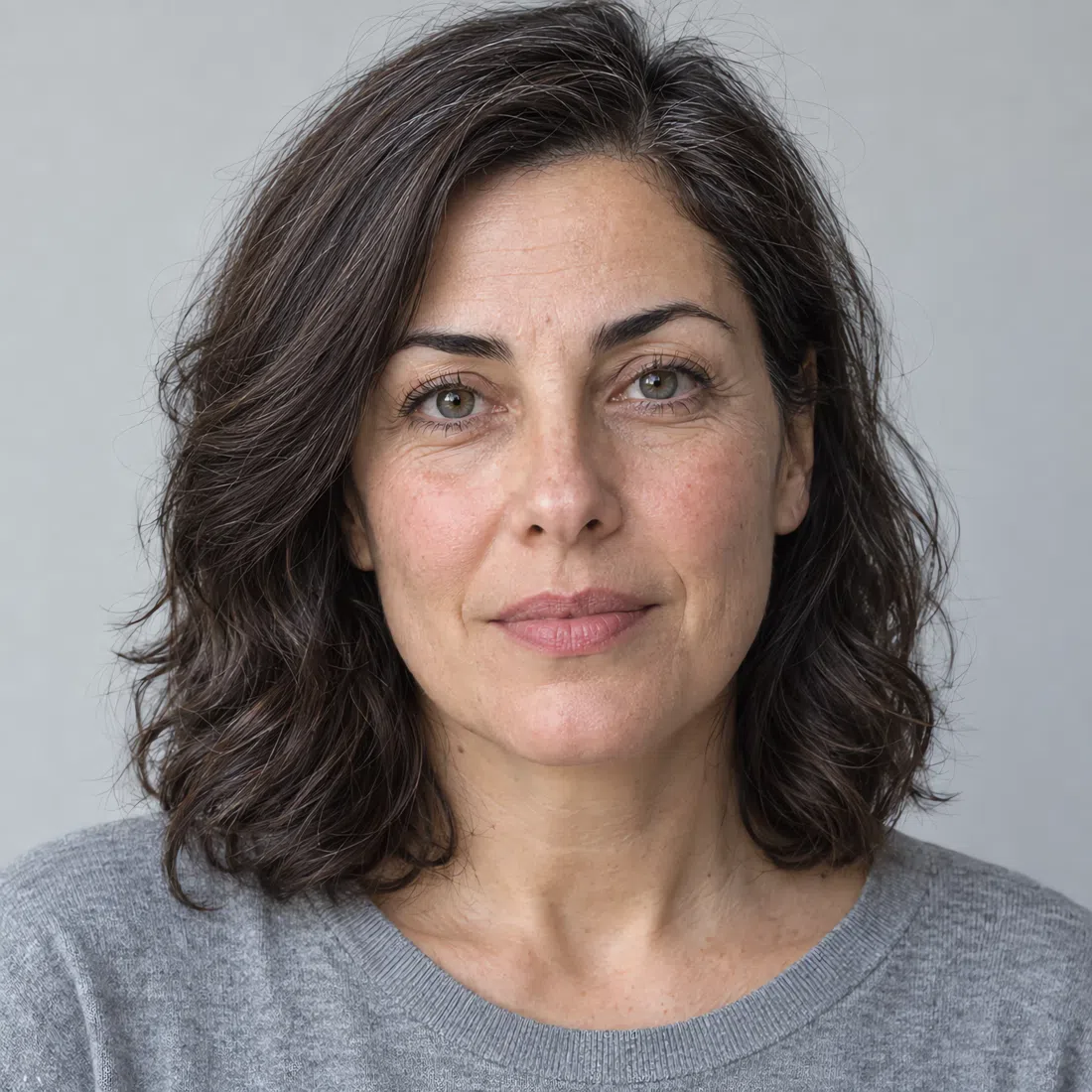 A Cool Summer woman with light olive skin, wavy dark ash brown hair and blue-gray eyes, in a plain gray top